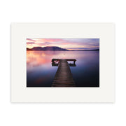 A wooden dock extends over Lake Tarawera toward distant mountains at sunset, framed with a white mat—Lake Tarawera - RP098, a stunning New Zealand photographic print by Reuben Price.