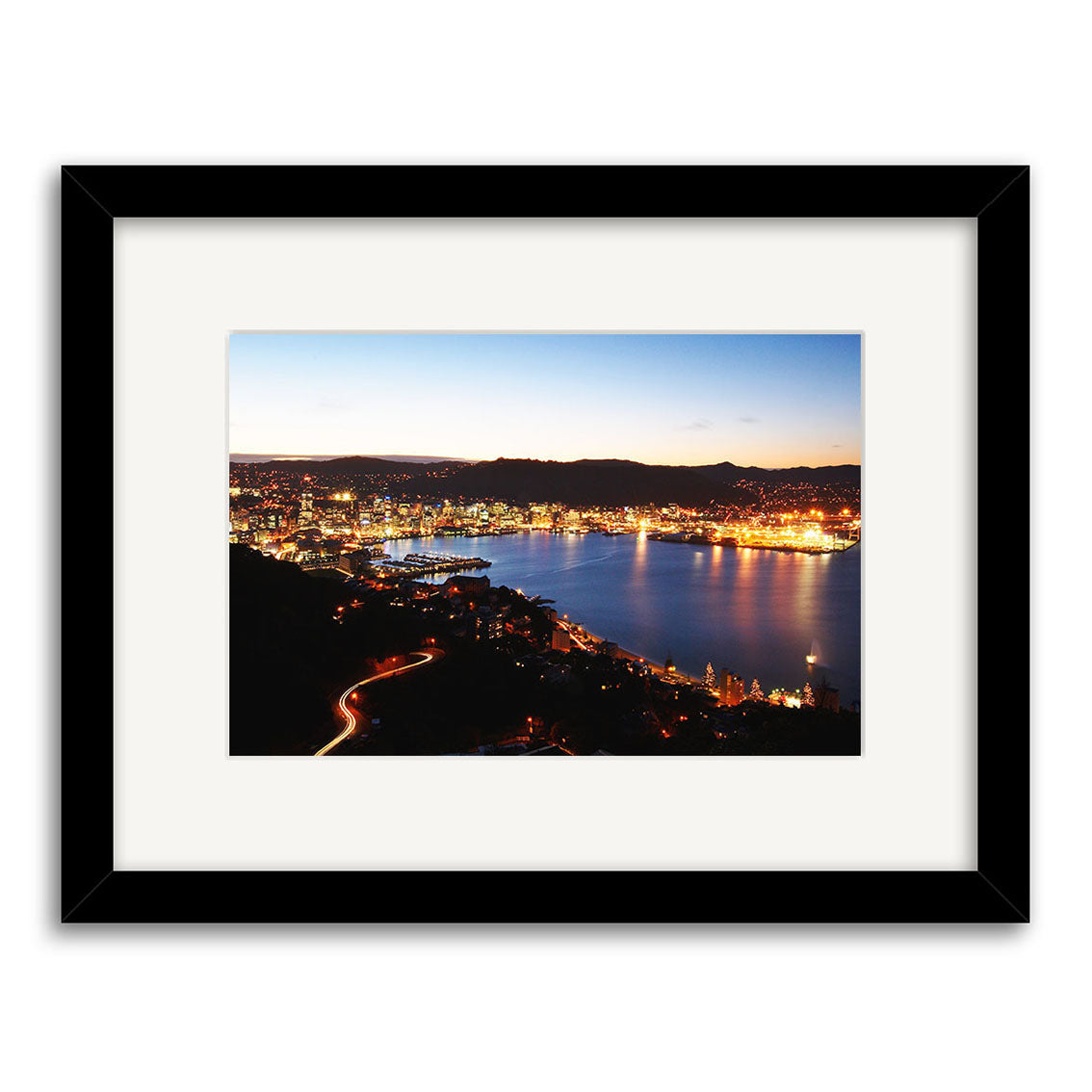 Framed photographic print, Wellington by Night - RP099, shows a New Zealand coastal city at dusk with city lights reflected on water, dark hills in back, and a light mat in a black frame. Photography by Reuben Price.