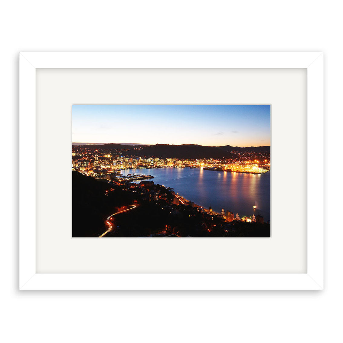 Wellington by Night - RP099 is a framed photographic print by Reuben Price showcasing Wellington’s waterfront at dusk, with city lights reflecting on the water. The white frame and wide white mat offer a modern finish.