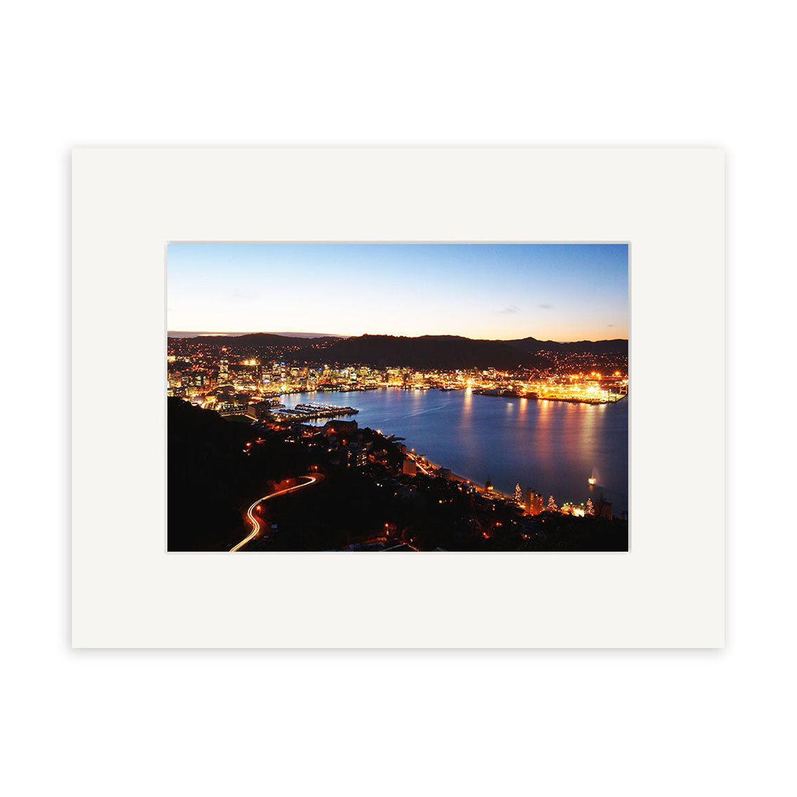 Wellington by Night - RP099, a photographic print by Reuben Price, captures waterfront buildings aglow at dusk in New Zealand with hills and car light trails, displayed with a white mat.