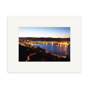 Wellington by Night - RP099, a photographic print by Reuben Price, captures waterfront buildings aglow at dusk in New Zealand with hills and car light trails, displayed with a white mat.