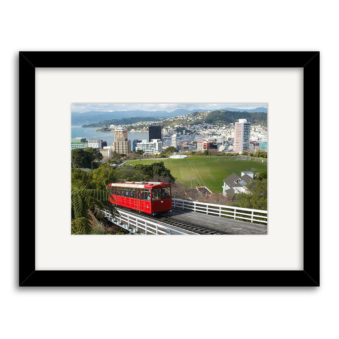 The Cable Car - RP100 shows a red cable car on a New Zealand track with cityscape, green hill, and mountains beneath a partly cloudy sky. Printed on archival matte paper, the image is framed with a black border and white mat.