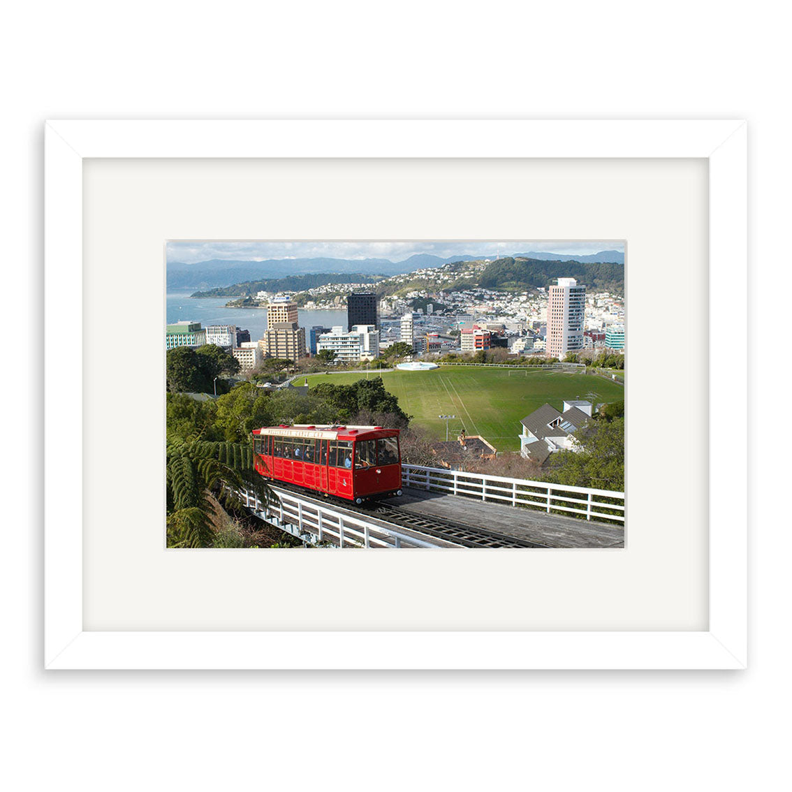 The Cable Car - RP100 print features a red cable car on a hillside track, overlooking a city, green fields, and distant hills. This New Zealand scene is framed in white and printed on archival quality matte paper.