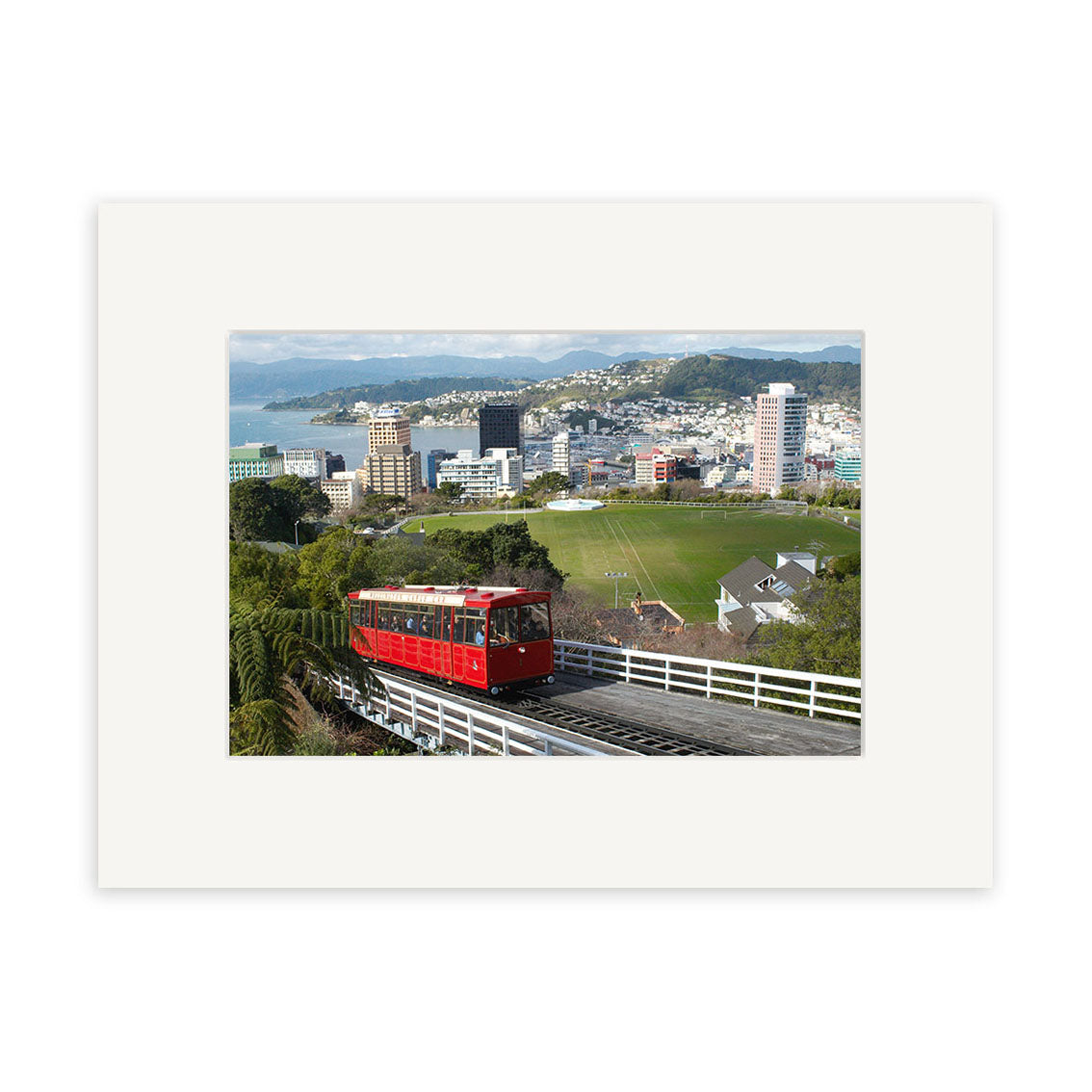 The Cable Car - RP100 features a red cable car ascending tracks in New Zealand, overlooking a city and greenery under a cloudy sky. Photographic prints on archival quality matte paper are available.