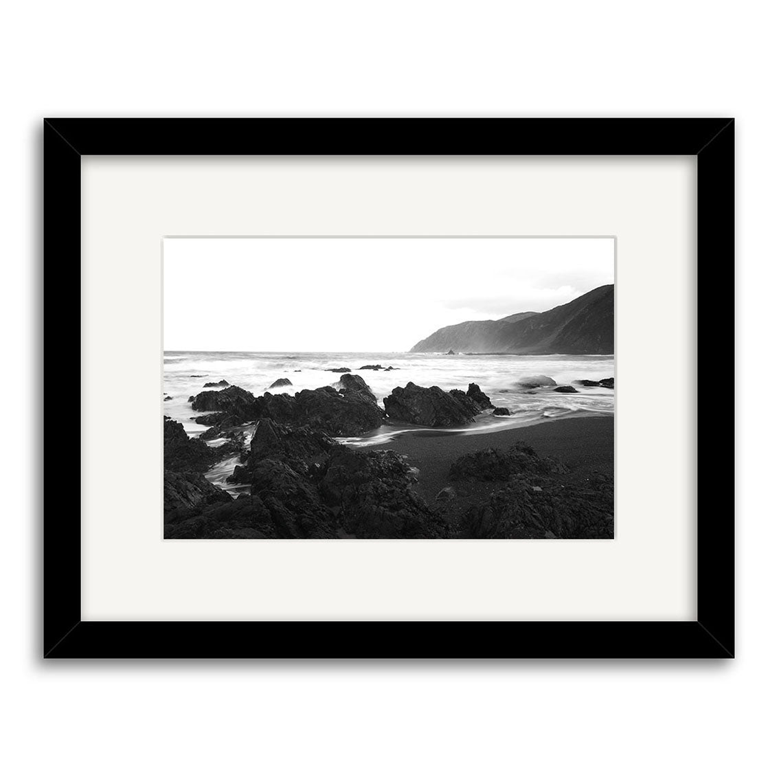 Owhiro Bay - RP102: Black and white photo by Reuben Price of waves crashing on a rocky New Zealand coastline, hills in the background. Presented as a photographic print in a simple black frame with white mat.
