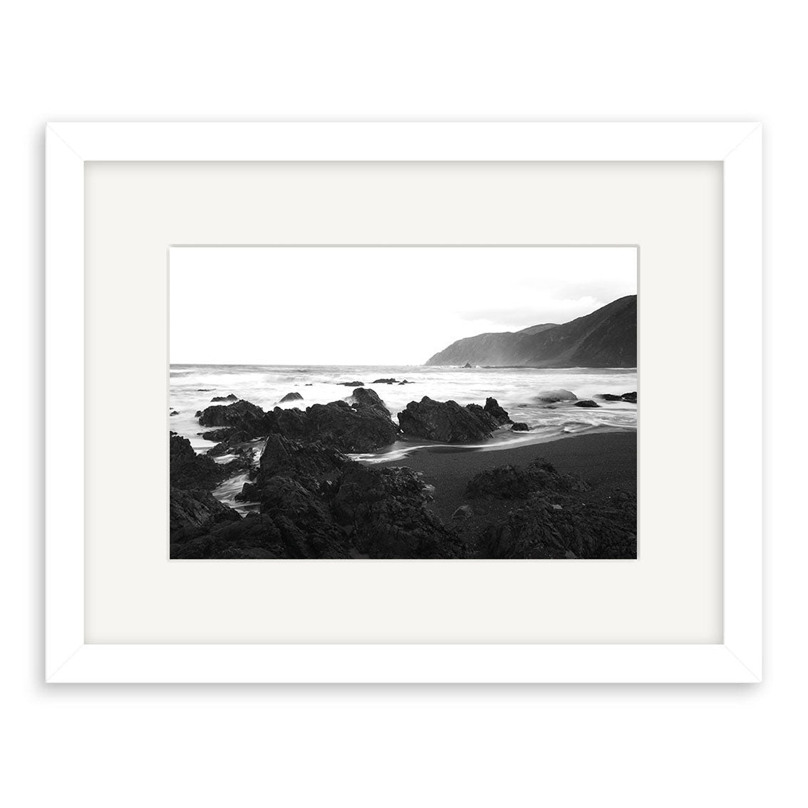 Owhiro Bay - RP102 is a black and white photographic print by Reuben Price, showcasing jagged rocks on a New Zealand shoreline with waves, distant hills, and a cloudy sky. It comes elegantly mounted in a white frame with a wide mat.