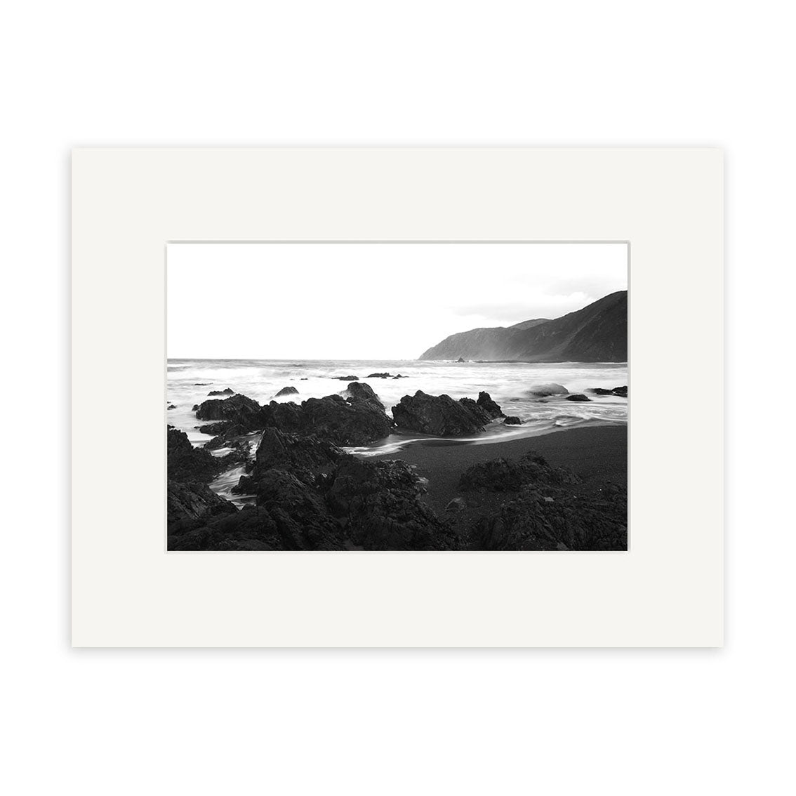 Owhiro Bay - RP102 is a black and white photo print featuring a rocky New Zealand shoreline with ocean waves, distant mountains, and a cloudy sky. Framed in a white mat.
