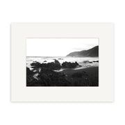 Owhiro Bay - RP102 is a black and white photo print featuring a rocky New Zealand shoreline with ocean waves, distant mountains, and a cloudy sky. Framed in a white mat.
