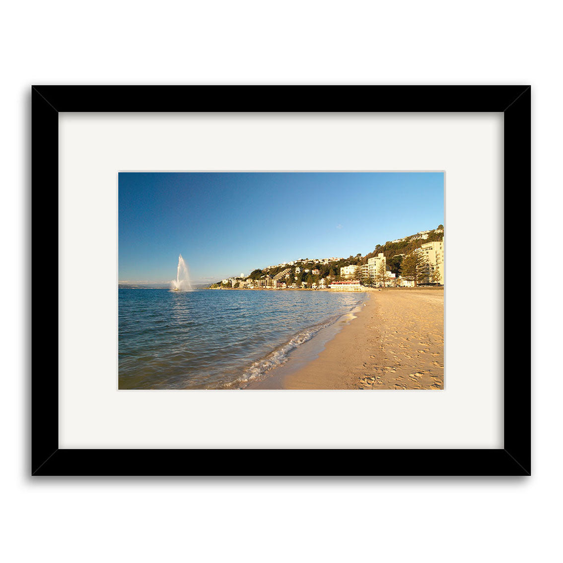 Oriental Bay - RP103: A framed archival matte print featuring a sandy New Zealand beach, shoreline buildings, gentle waves, and a sea fountain beneath a clear blue sky.