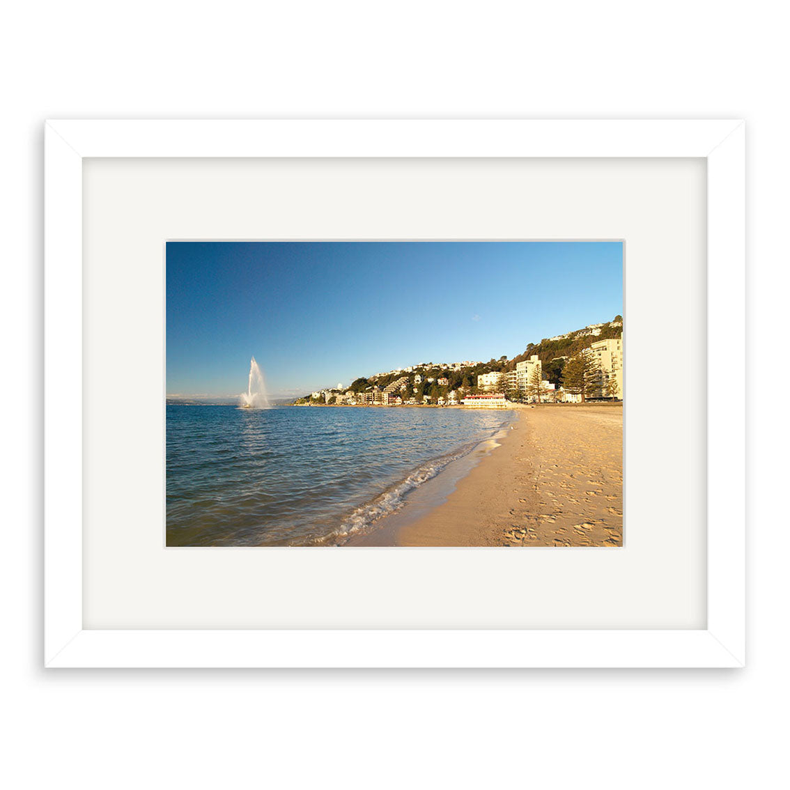 A photograph titled Oriental Bay - RP103 shows a sandy beach with gentle waves, shoreline buildings, and a large sea fountain, beautifully framed with a white matte and printed on archival quality matte paper.