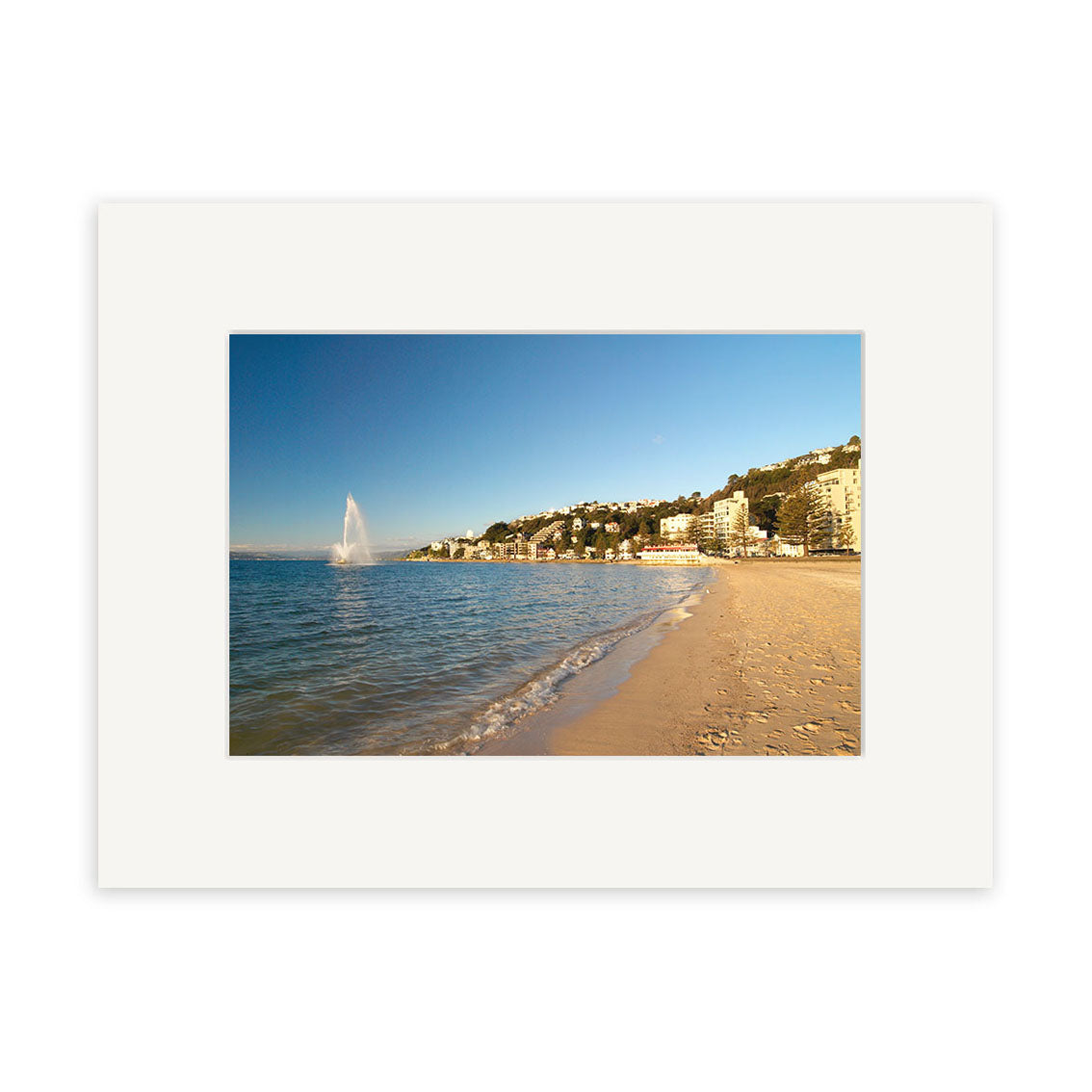 Experience the coastal beauty of New Zealand with Oriental Bay - RP103—a photographic print featuring a sandy beach, city skyline, and a dramatic water fountain under clear blue skies.