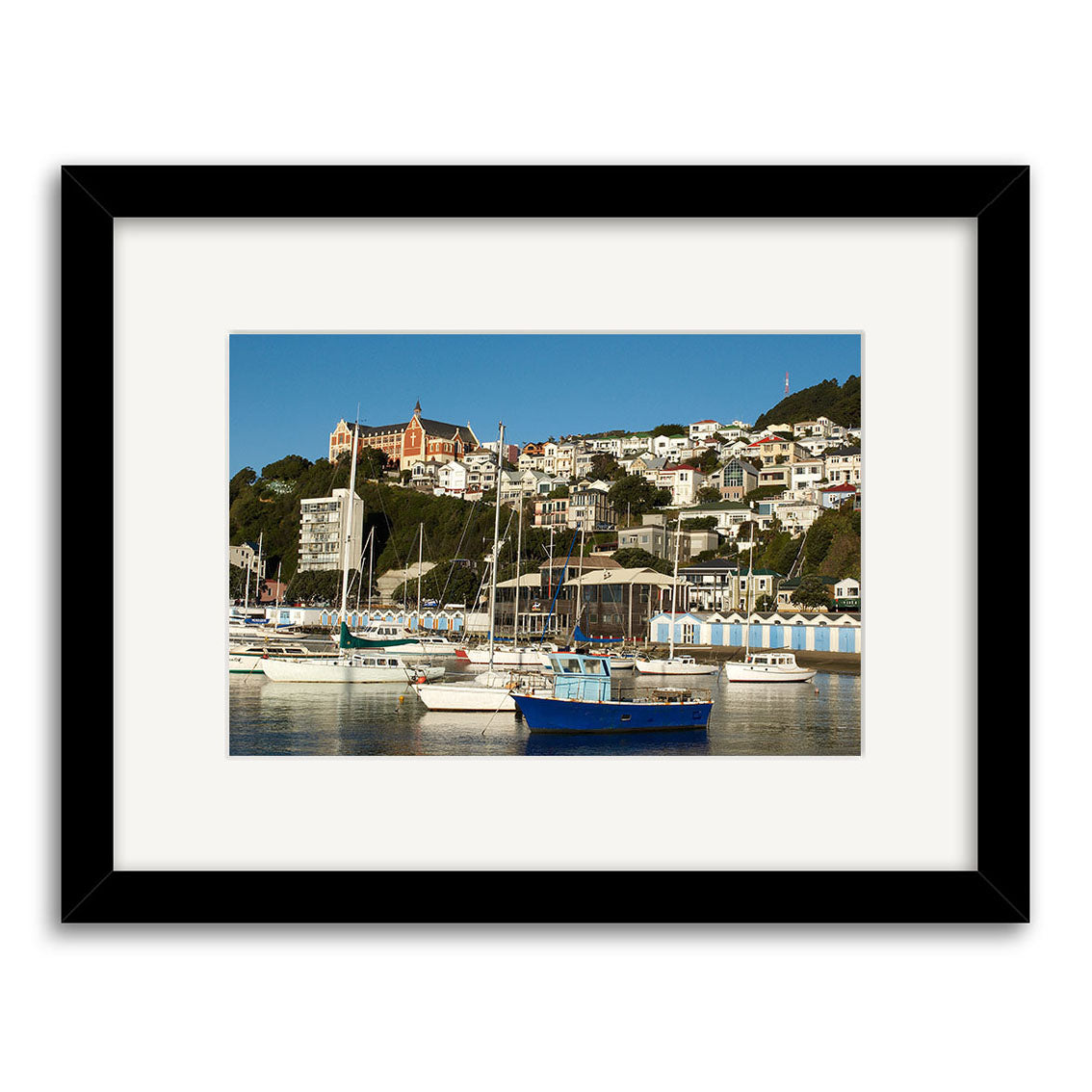 Oriental Bay Boats - RP104: Framed photographic print featuring boats in a New Zealand marina, white hillside houses, a red-roofed building, and clear blue sky. Printed on archival quality paper.