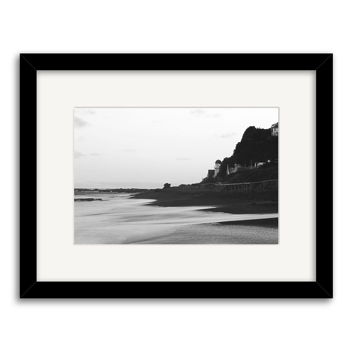 Island Bay - RP106: Black and white framed photo of a tranquil beach, calm water, sandy shore, and distant hill with structures—including a domed building—under clear skies. Printed on archival quality matte paper.