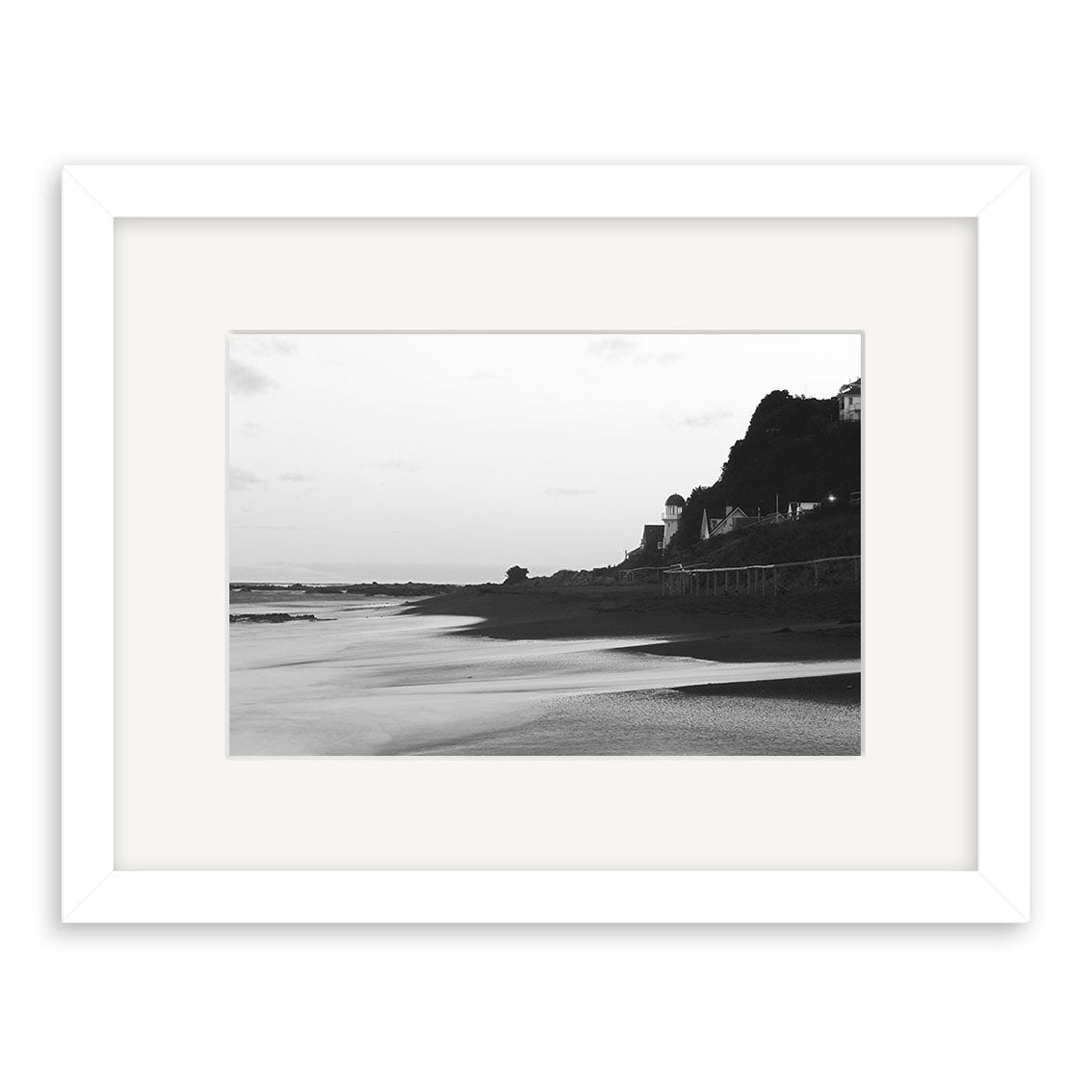 Island Bay - RP106: Black and white photo of a beach at low tide with calm water, distant shoreline, and hillside buildings. Displayed in a white mat and frame. Printed on archival quality matte paper by Reuben Price.