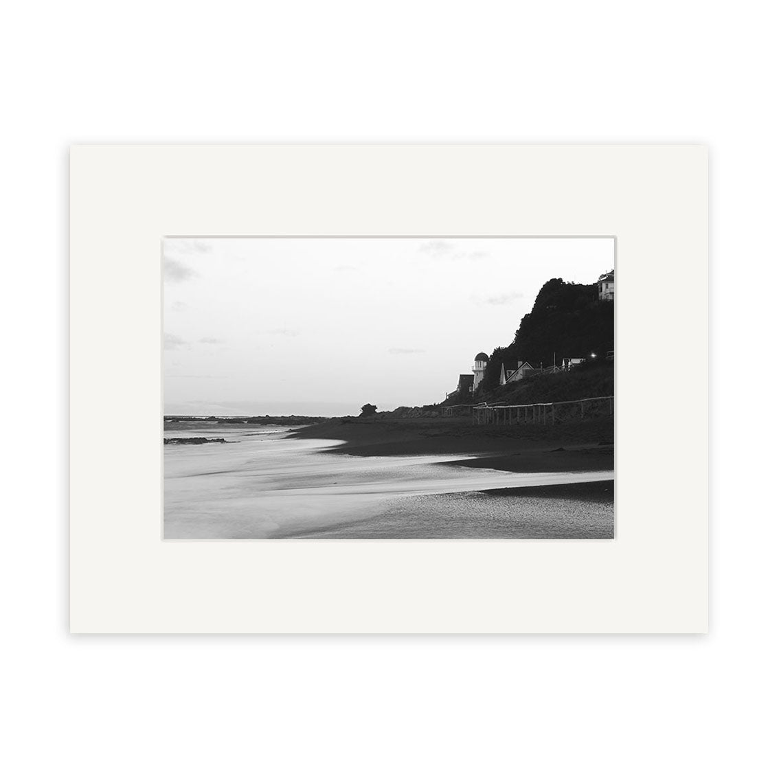 Island Bay - RP106 features a black and white photo of smooth sand, calm water, hillside buildings, and a pier—bordered by a white mat frame. Printed on archival matte paper, it captures Reuben Price’s classic New Zealand style.