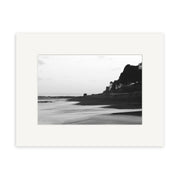 Island Bay - RP106 features a black and white photo of smooth sand, calm water, hillside buildings, and a pier—bordered by a white mat frame. Printed on archival matte paper, it captures Reuben Price’s classic New Zealand style.