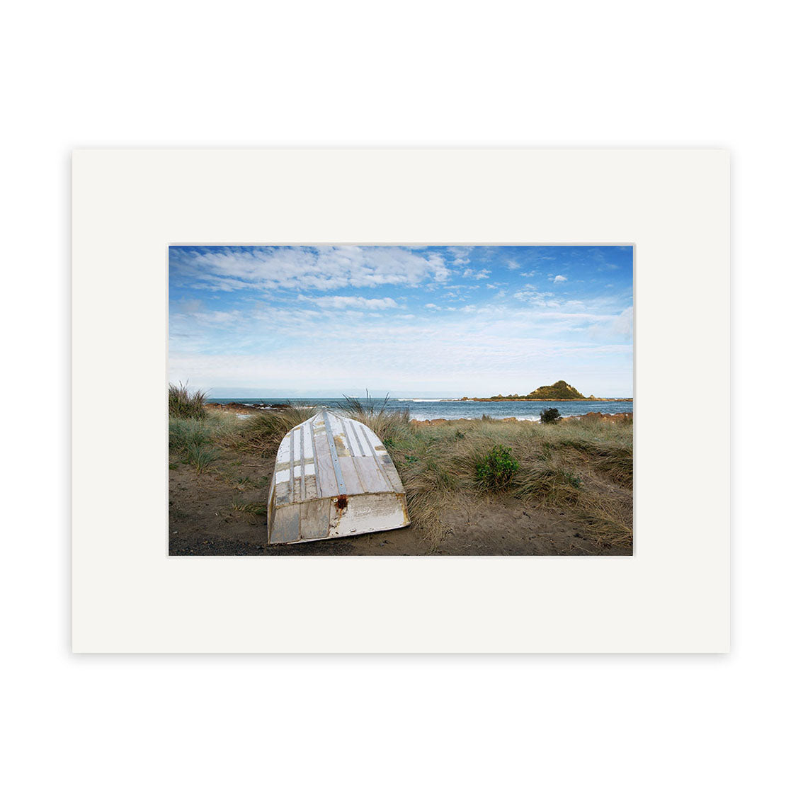The photographic print Island Bay 2 - RP107 by Reuben Price depicts a white wooden rowboat flipped on sandy grass, with a beach, ocean, and distant rocky island under a partly cloudy New Zealand sky.