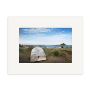 The photographic print Island Bay 2 - RP107 by Reuben Price depicts a white wooden rowboat flipped on sandy grass, with a beach, ocean, and distant rocky island under a partly cloudy New Zealand sky.