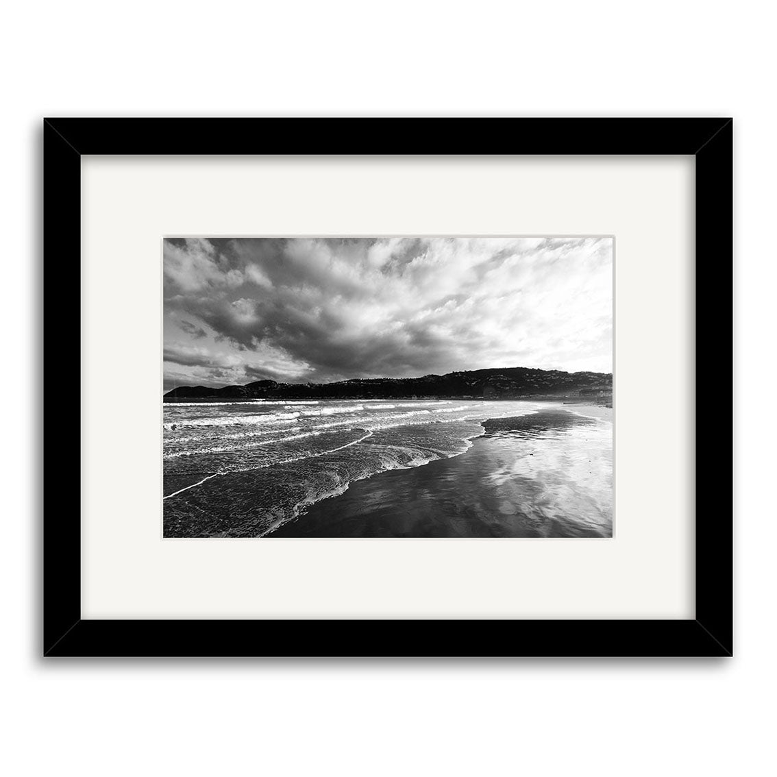 Lyall Bay - RP108: Black-and-white photo of waves on a New Zealand beach with distant hills under a cloudy sky, framed with a white mat and black frame. Photographic print on archival matte paper for lasting beauty.
