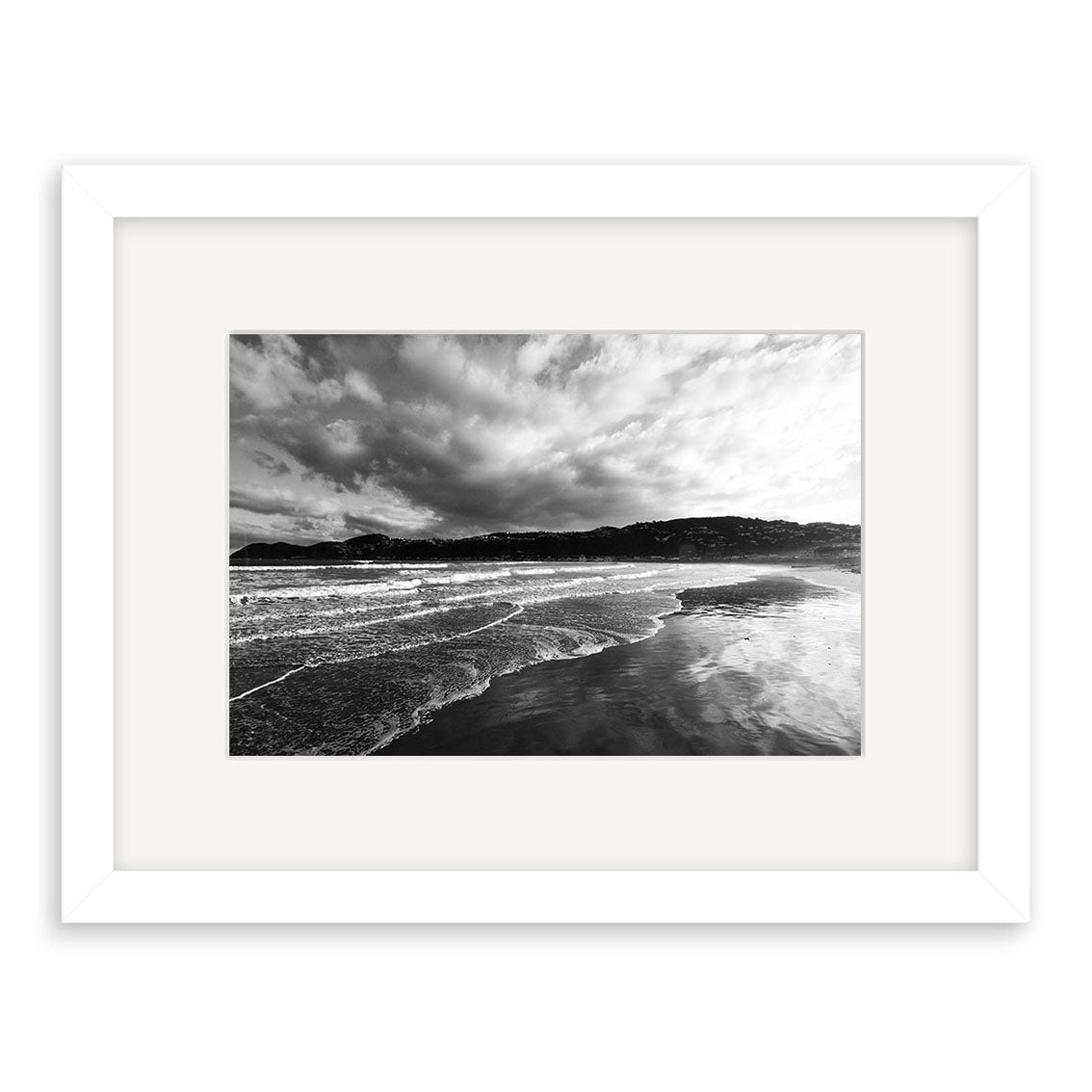 Lyall Bay - RP108: A framed black and white photograph capturing ocean waves on a sandy New Zealand beach, with distant hills and dramatic clouds. Available as archival quality matte paper photographic prints.