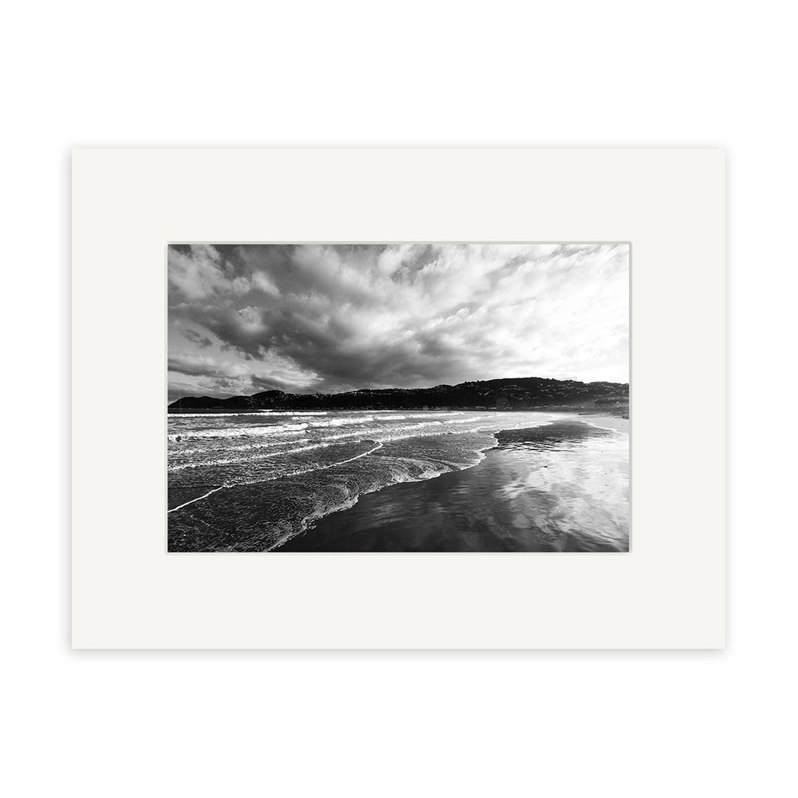 Lyall Bay - RP108: A black and white photo of ocean waves on a sandy New Zealand beach beneath cloudy skies, distant hills visible. Printed on archival matte paper and presented with a white mat board.