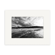 Lyall Bay - RP108: A black and white photo of ocean waves on a sandy New Zealand beach beneath cloudy skies, distant hills visible. Printed on archival matte paper and presented with a white mat board.