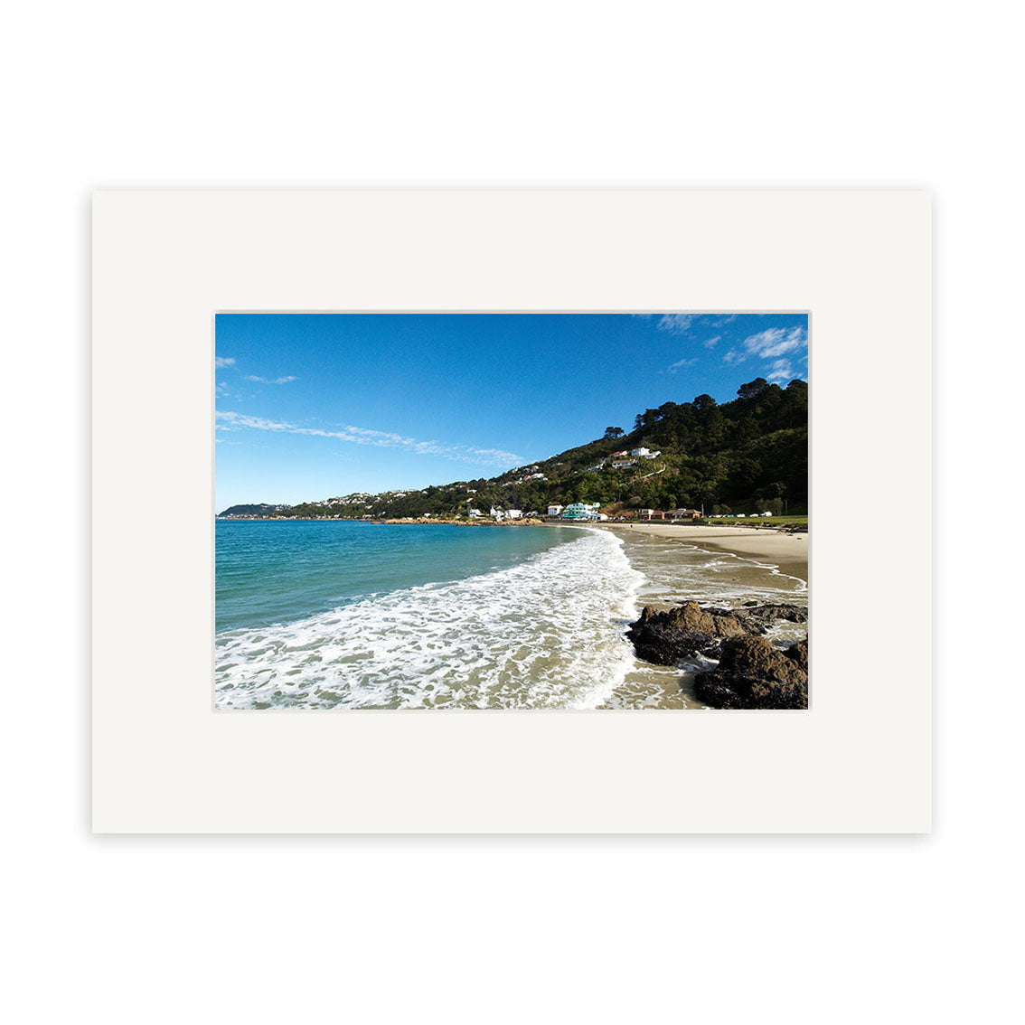 “Scorching Bay - RP109: A photo of a New Zealand beach with gentle waves, sandy shore, hillside houses and greenery beneath a clear blue sky. Presented in a white mat frame on archival quality matte paper.”.