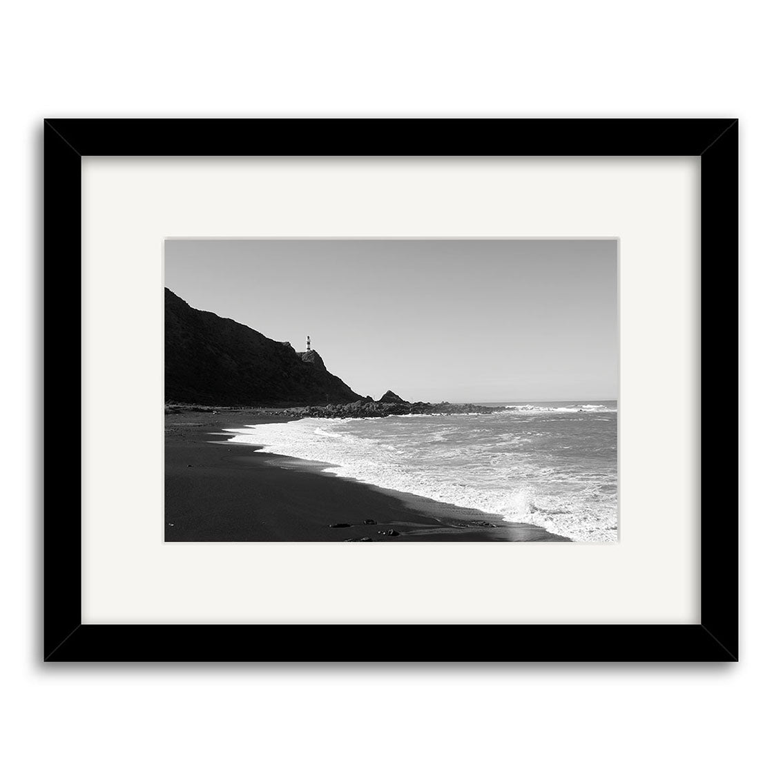 Cape Palliser - RP111: Black and white photograph of a New Zealand coastal scene featuring waves on a sandy beach and a lighthouse on a rocky hill, presented in an archival white mat with black frame.