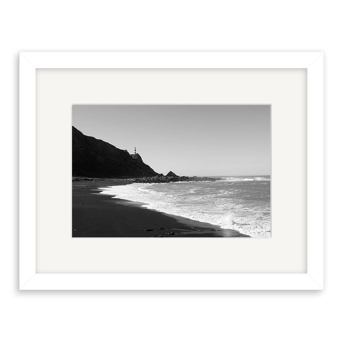Cape Palliser - RP111 is an archival quality black and white photo print of a New Zealand beach with gentle waves, a rocky coastline, and a lighthouse on a hill, elegantly presented in a white mat and frame.