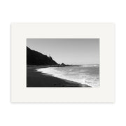 Cape Palliser - RP111: Black and white photo of a New Zealand beach with waves, rocky cliffs, and a distant lighthouse under clear sky, framed by a white mat—perfect for archival-quality photographic prints.