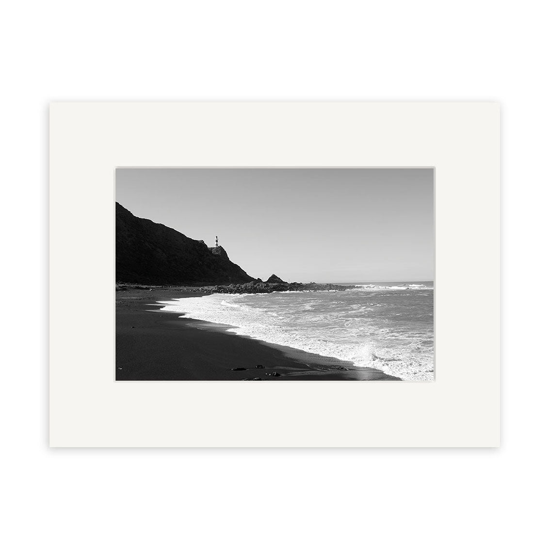 Cape Palliser - RP111: Black and white photo of a New Zealand beach with waves, rocky cliffs, and a distant lighthouse under clear sky, framed by a white mat—perfect for archival-quality photographic prints.