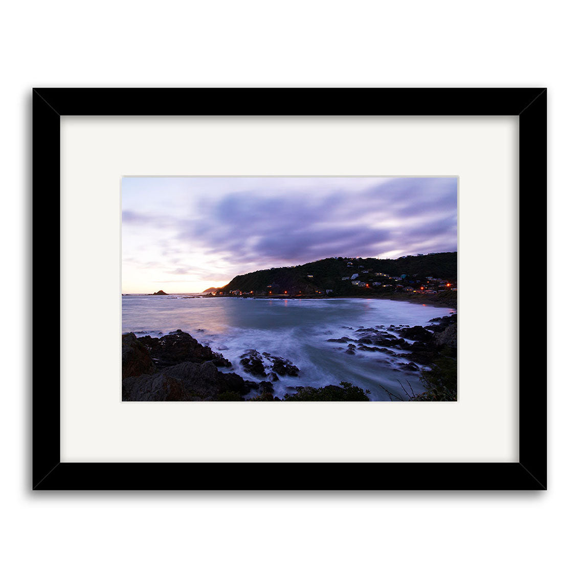 Houghton Bay - RP112 is a framed, archival-quality photographic print by Reuben Price, depicting a rocky shoreline, calm waves, hillside homes, and a cloud-filled dusk sky illuminated by the fading sun.