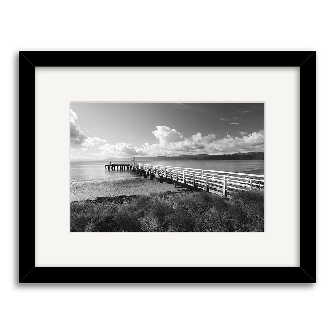 Seatoun - RP113: A black and white photo of a wooden pier in New Zealand stretches over calm water under a cloudy sky, with grassy foreground. Framed with a white mat and black frame on archival quality matte paper.