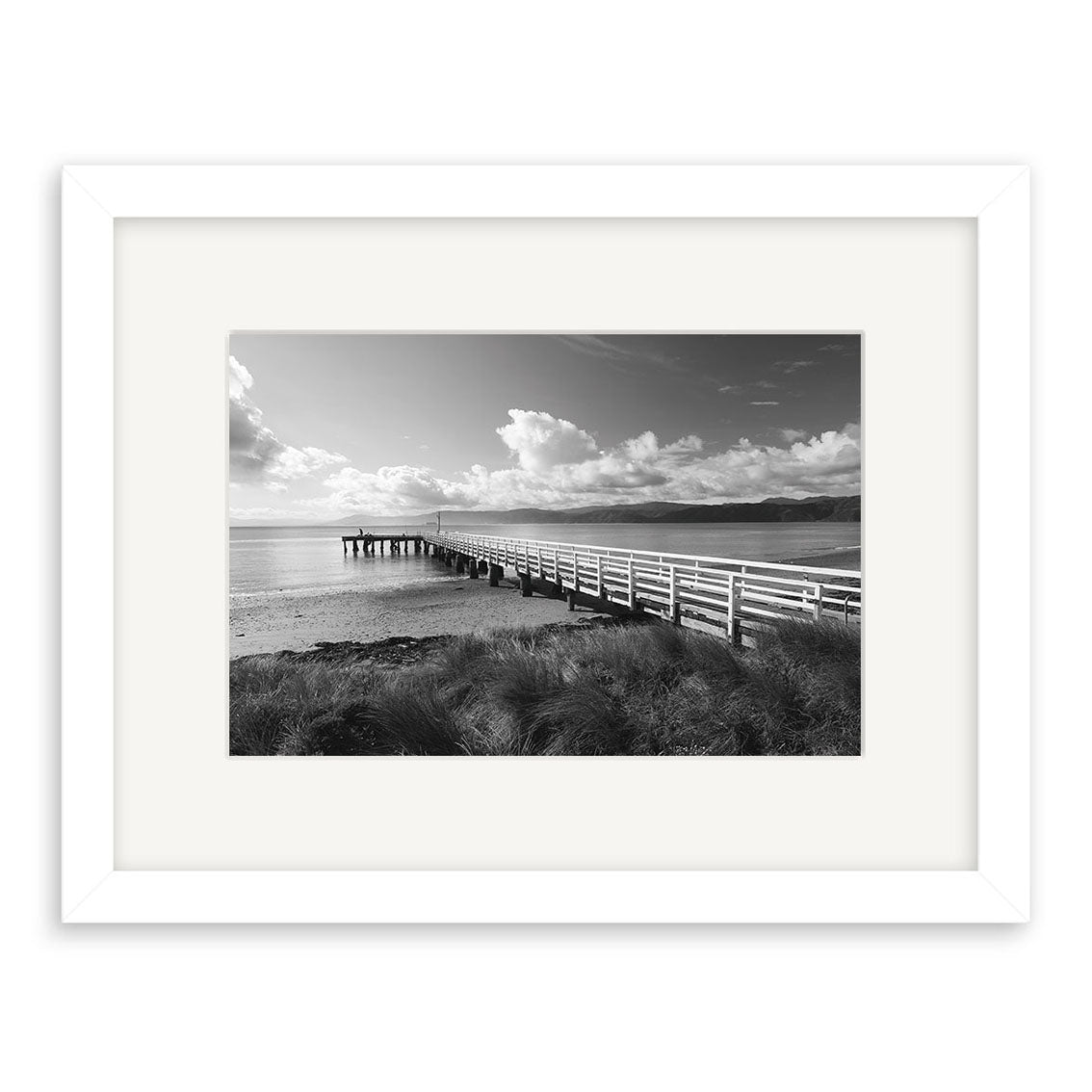 Seatoun - RP113: Black and white photo of a long wooden pier over water with grassy dunes, partly cloudy skies, framed with a white mat. Available as archival quality matte paper photographic prints.