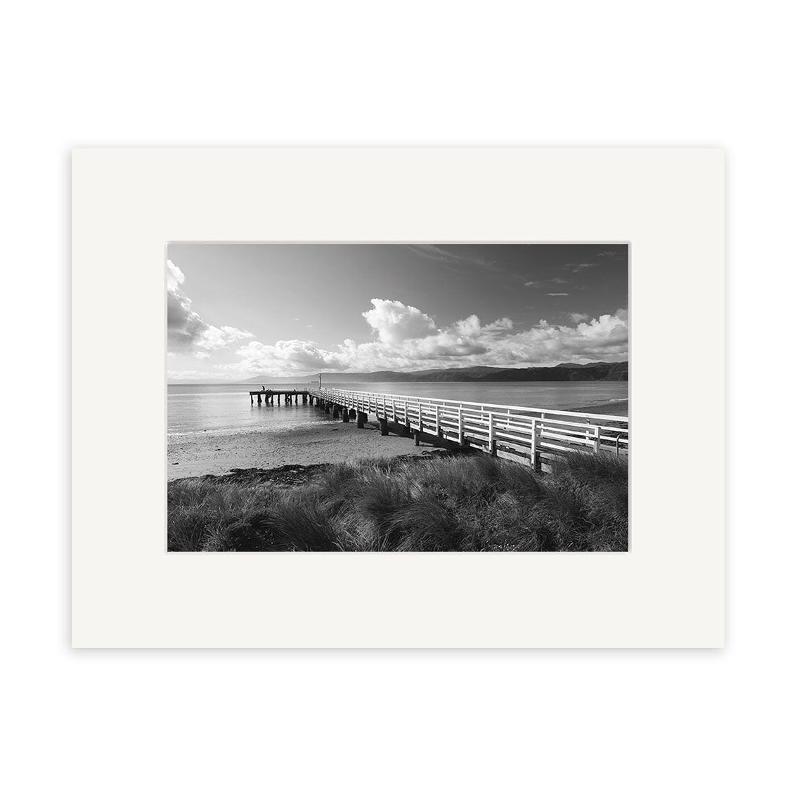 The Seatoun - RP113 is a black and white photo of a wooden pier over calm water, with grass and distant hills under a cloudy sky. Printed on archival matte paper, this photographic print comes framed with a white mat.