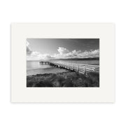 The Seatoun - RP113 is a black and white photo of a wooden pier over calm water, with grass and distant hills under a cloudy sky. Printed on archival matte paper, this photographic print comes framed with a white mat.