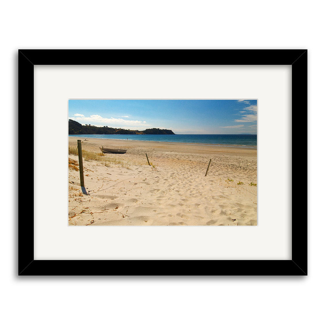 Onetangi - Waiheke Island - RP116: A framed photographic print of a sandy New Zealand beach with a small boat, gentle waves, and a headland under blue sky, in a black frame with white mat on archival matte paper.