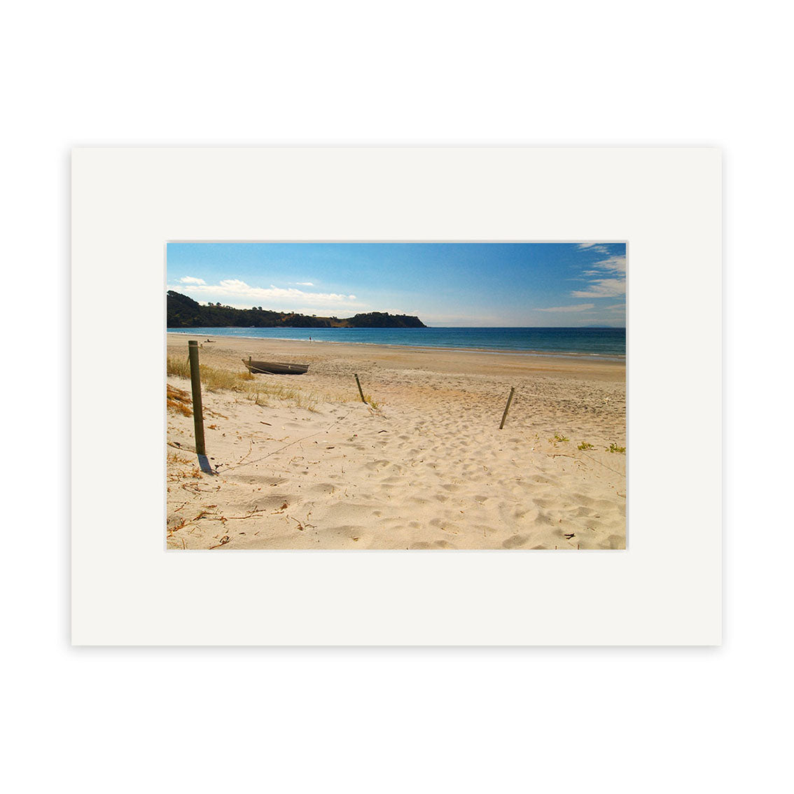 “Onetangi - Waiheke Island - RP116” features a small boat on a sandy New Zealand beach, grassy dunes, calm sea, and distant headland under a partly cloudy sky. Available as archival quality photographic prints with white mat frame.