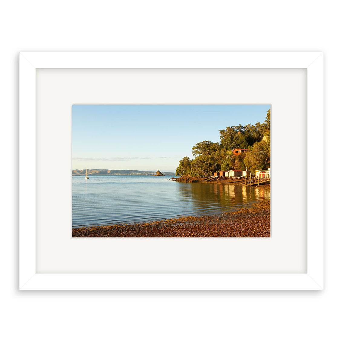 A framed photographic print, Rocky Bay - RP118 by Reuben Price, featuring a calm New Zealand shoreline with pebbled beach, still water, green trees, and small coastal buildings under a clear blue sky.