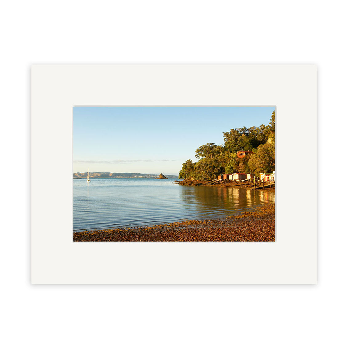 Experience the tranquility of New Zealand’s Rocky Bay in Rocky Bay - RP118, a stunning photographic print by Reuben Price featuring clear waters, a pebbled beach, green trees, scattered buildings, and distant land under a clear sky.