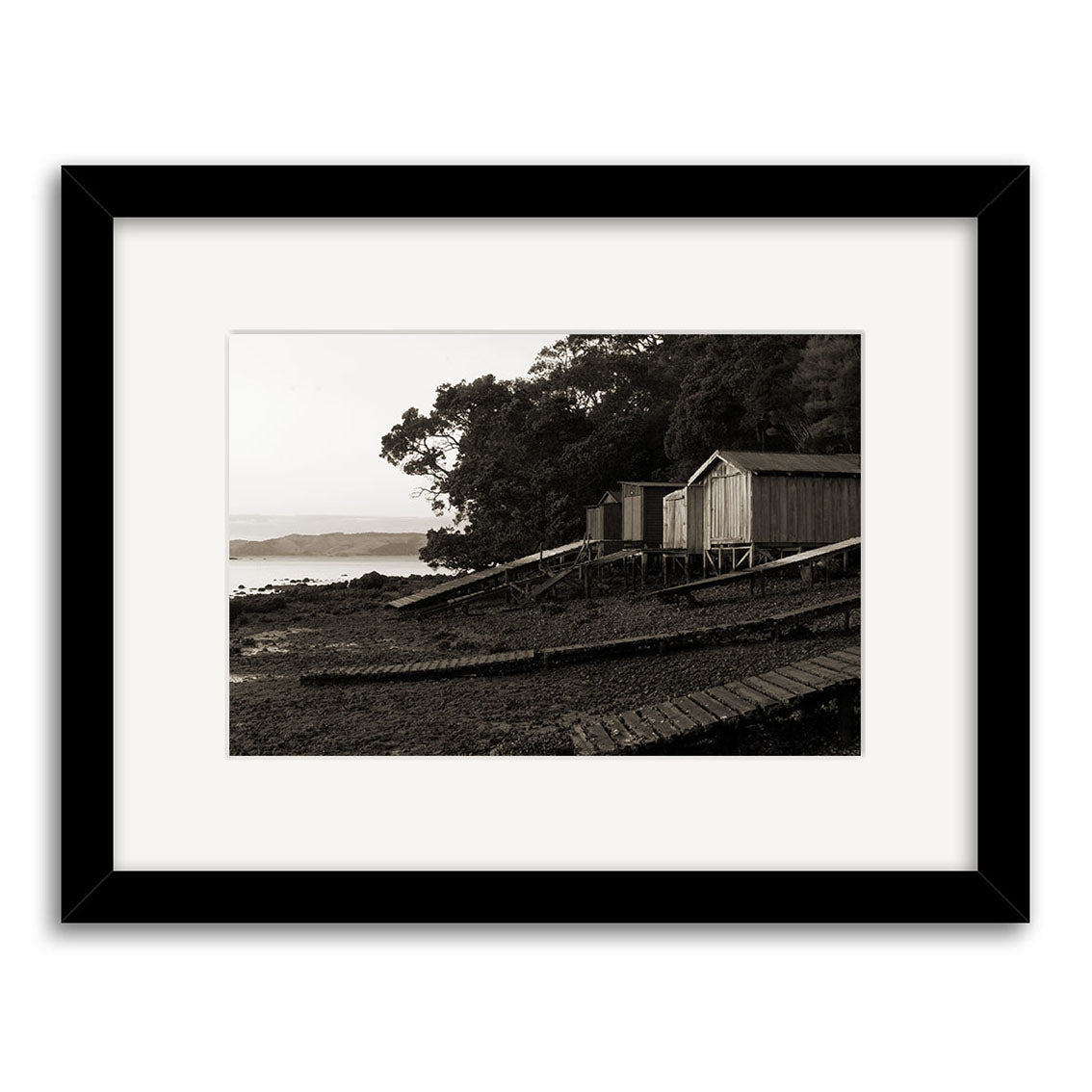 Rocky Bay - RP119 is a black and white photo of three wooden sheds on stilts by a rocky New Zealand shore, shown in a black frame with white matting, printed on archival quality matte paper.