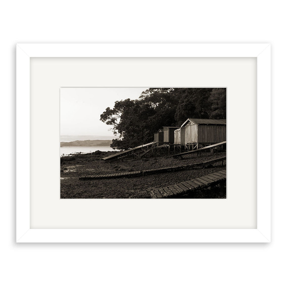 The black-and-white photo Rocky Bay - RP119 features wooden boathouses on stilts by a rocky New Zealand shoreline, surrounded by trees and ramps—available as archival quality matte paper photographic prints.