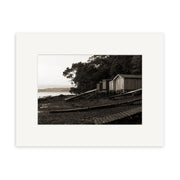 Rocky Bay - RP119 is a black and white photo of three wooden sheds on stilts by a rocky New Zealand shore with tree foliage behind and water in the background, framed with a white mat and printed on archival matte paper.