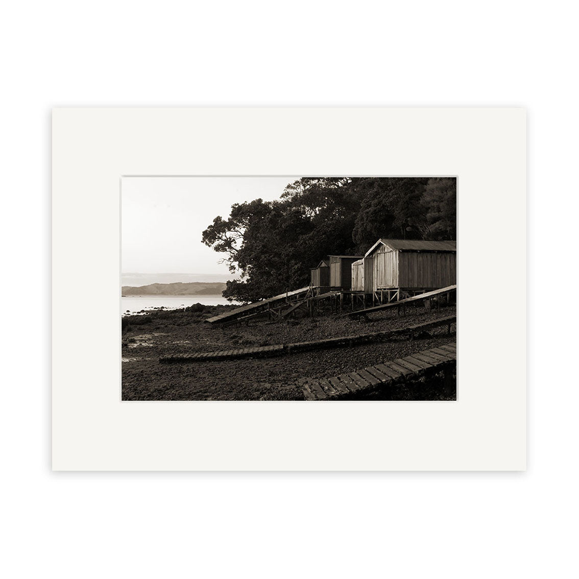 Rocky Bay - RP119 is a black and white photo of three wooden sheds on stilts by a rocky New Zealand shore with tree foliage behind and water in the background, framed with a white mat and printed on archival matte paper.
