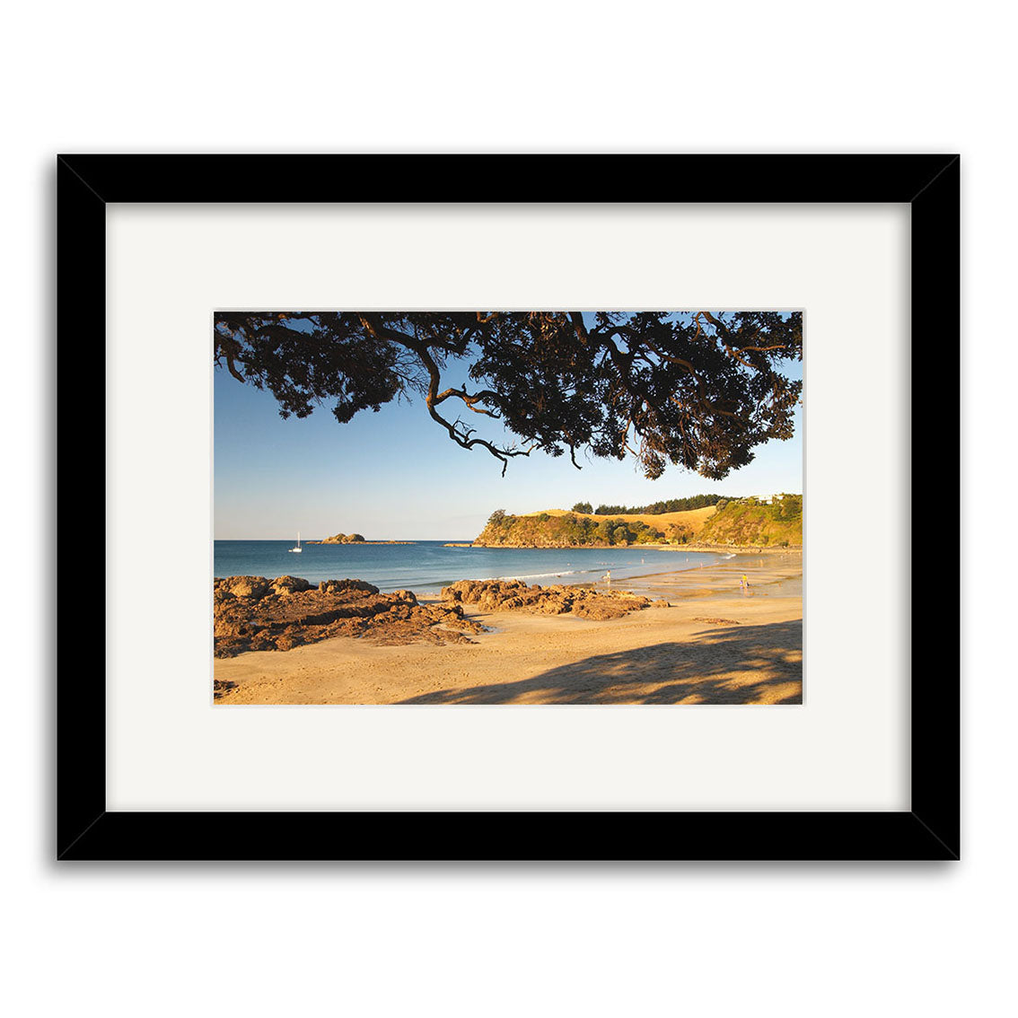 Introducing Palm Beach - Waiheke - RP120: a framed photographic print featuring a sandy New Zealand beach with rocks, calm blue water, distant hills, sailboat, and tree branches. Printed on archival quality paper.