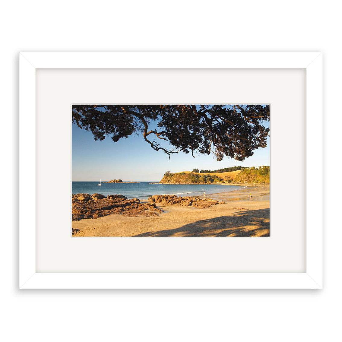 Palm Beach - Waiheke - RP120: An archival photographic print features a sandy New Zealand beach with rocks, calm blue water, a distant sailboat, tree branch shadows, and green hills on the horizon under a clear sky.