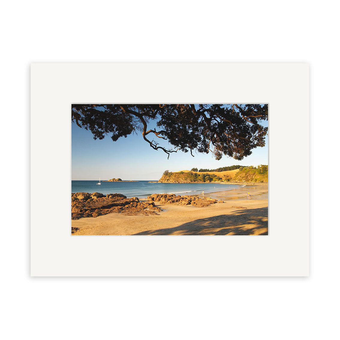 Palm Beach - Waiheke - RP120 features a sandy beach with rock formations, tranquil blue water, a distant sailboat, leafy trees, and grassy hills—classic New Zealand scenery perfect for archival quality photographic prints.