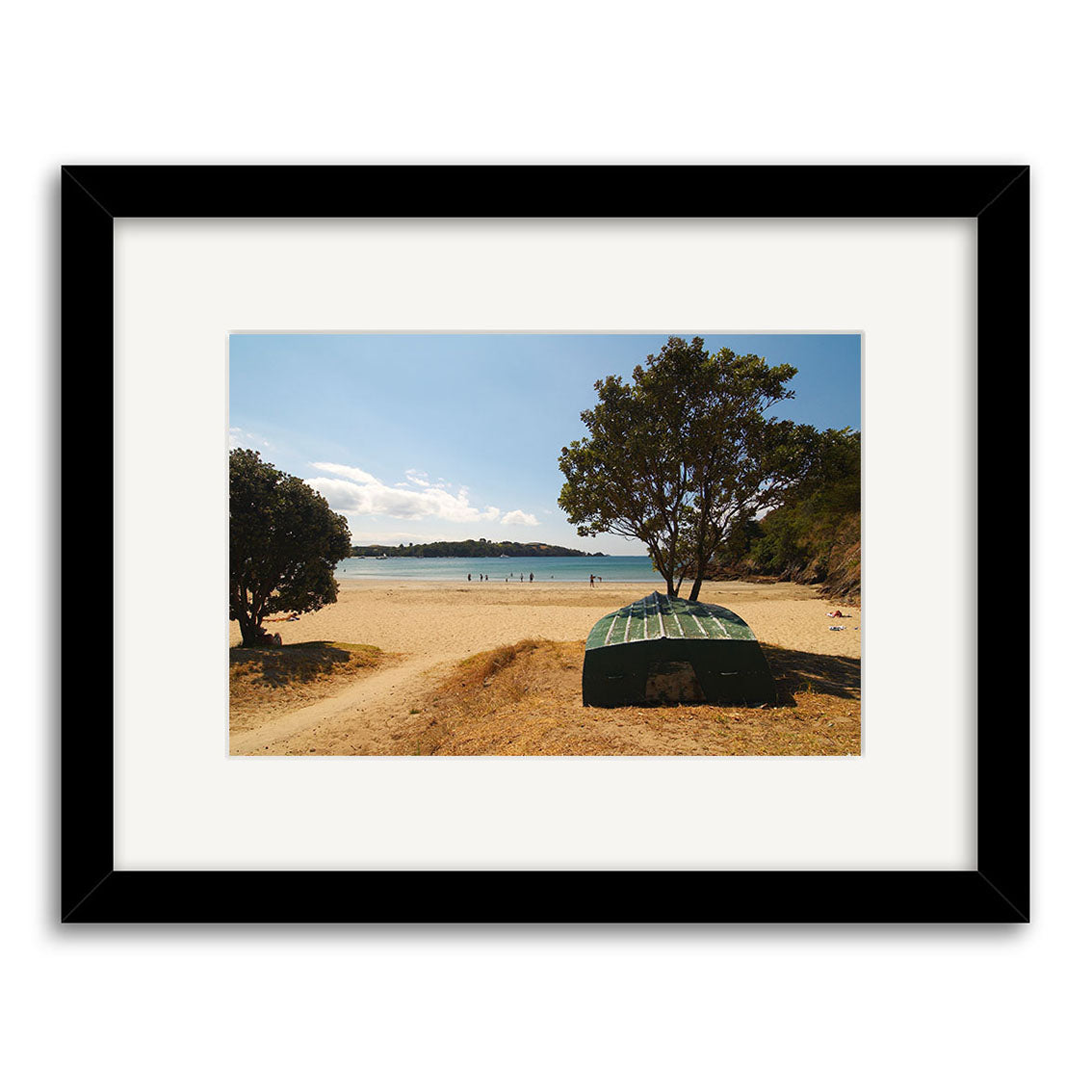 The Little Oneroa - Waiheke - RP121 framed photographic print captures a green boat overturned by two trees on a sandy New Zealand beach, with people in the distance near the shoreline under a clear blue sky.