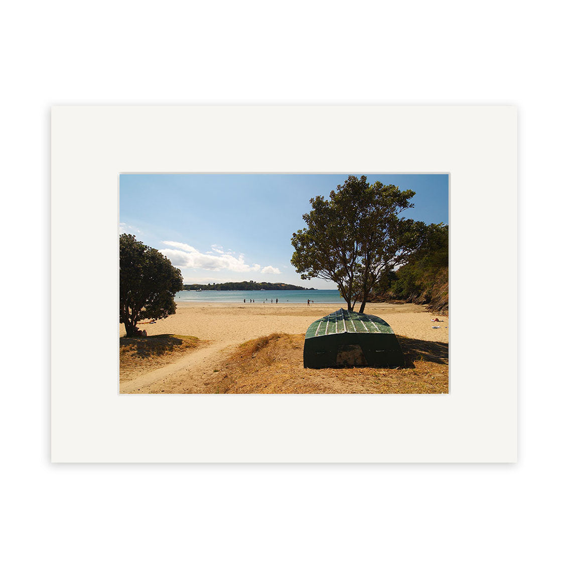 The photographic print Little Oneroa - Waiheke - RP121 by Reuben Price features a small green boat upside down on grass beneath a tree near a sandy New Zealand beach, with people on the shore and sparkling blue water under a sunny sky.