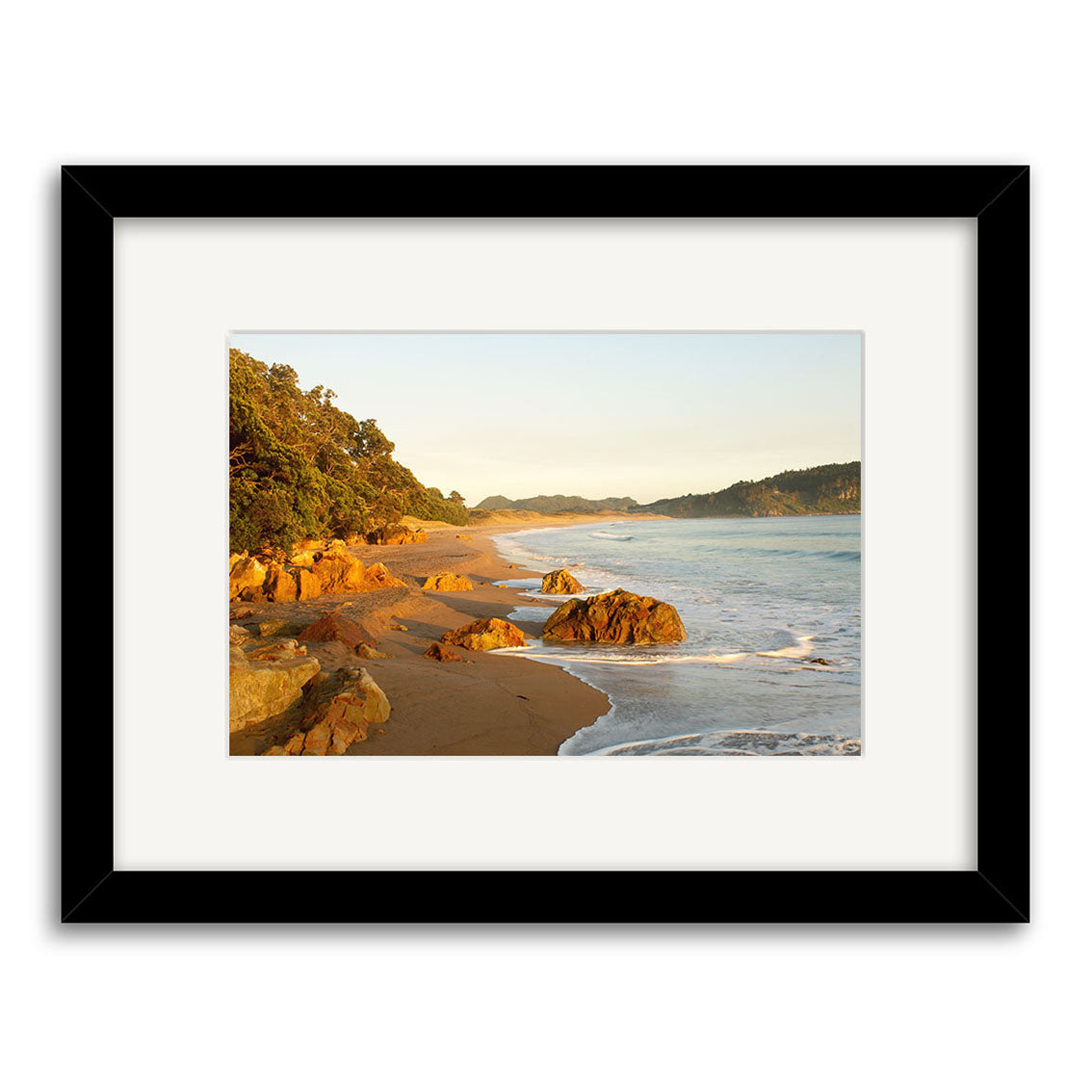 Hot Water Beach - RP122 is a framed photograph featuring sandy shores, ocean waves, rocks, and lush green trees. This Reuben Price print is crafted on archival-quality matte paper for lasting beauty and displayed against a white background.